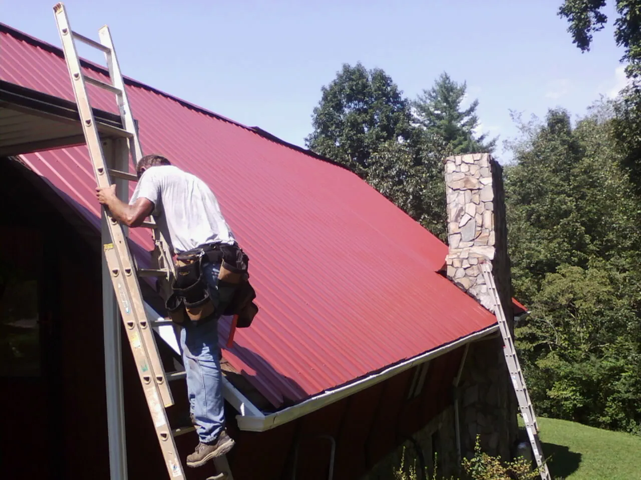 Roofer on a ladder beside a red metal roof and stone chimney — Soffit & Fascia Repair by Roofing Brothers in Robersonville
